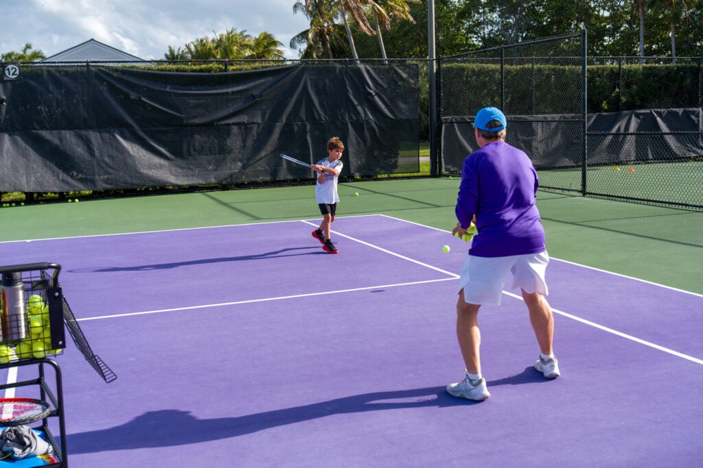 A child practices hitting a tennis ball with a racket while an adult observes on a purple outdoor tennis court.