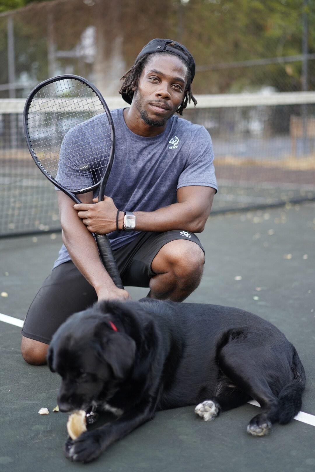 A man in athletic wear kneels on a tennis court holding a racket, while a black dog lies in front of him chewing on an object.