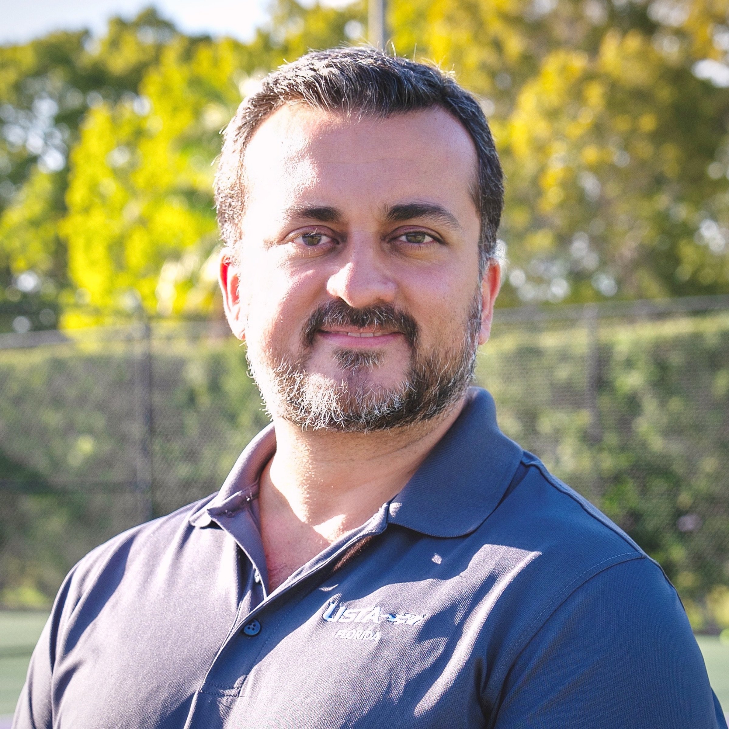 A man with short dark hair and a beard stands outdoors in front of a tennis court, wearing a dark polo shirt. Trees and a fence are visible in the background.