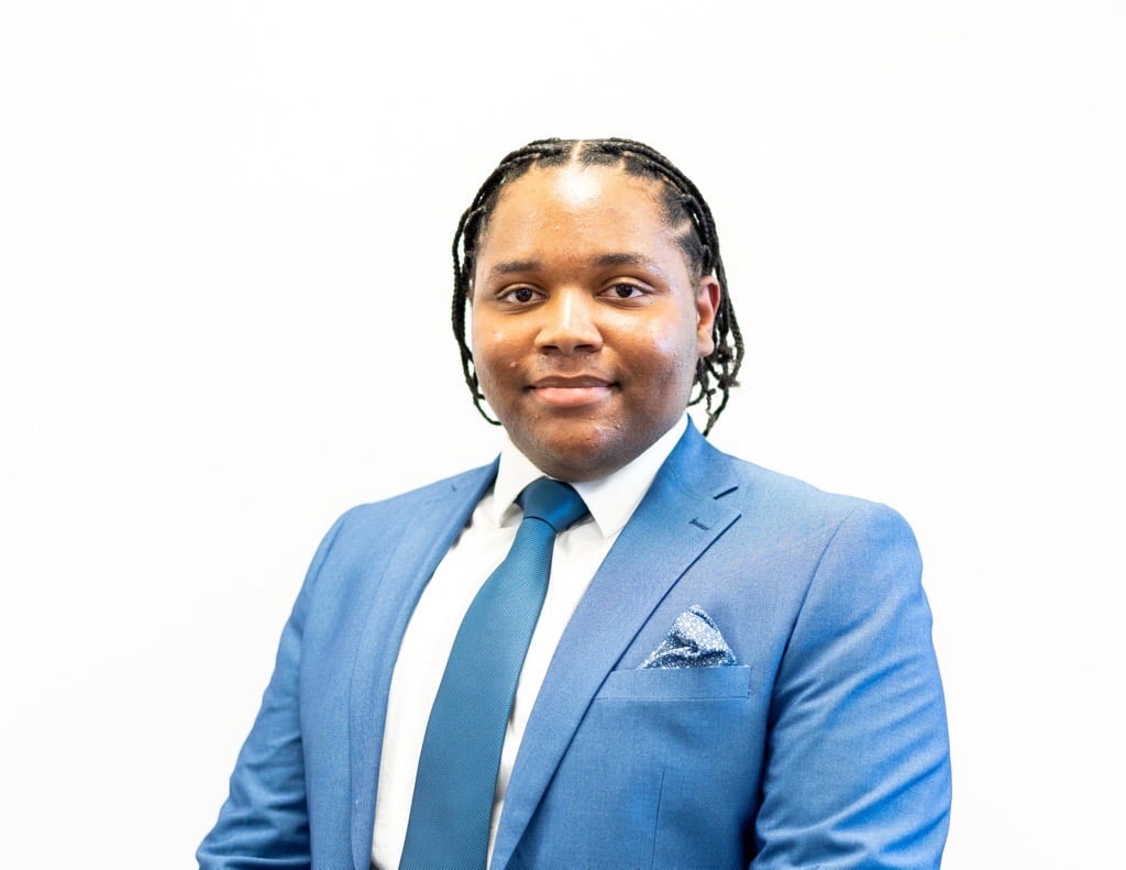 A person in a blue suit, white shirt, and blue tie poses against a plain white background.