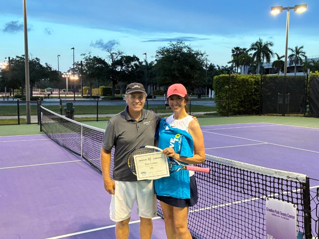 Two people stand on a purple tennis court; one holds a certificate and a gift bag. They are smiling and posing near the net with tennis rackets. Palm trees and lights are in the background.