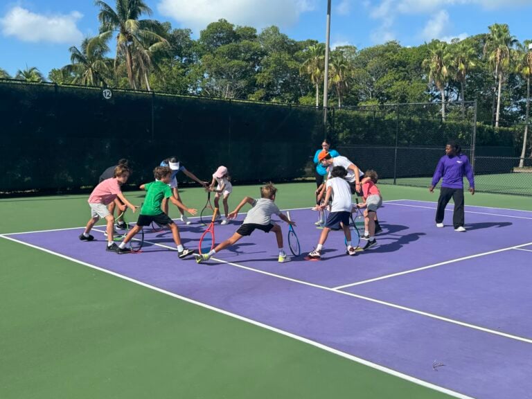 A group of children, each holding a tennis racket, gather in a circle on a purple tennis court while two adults supervise.