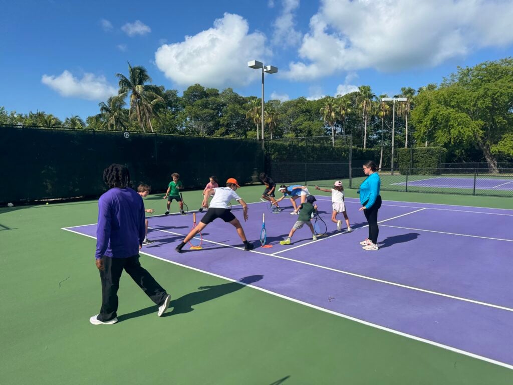 A group of children and two adults are on a purple tennis court, participating in an outdoor tennis lesson under a sunny sky.