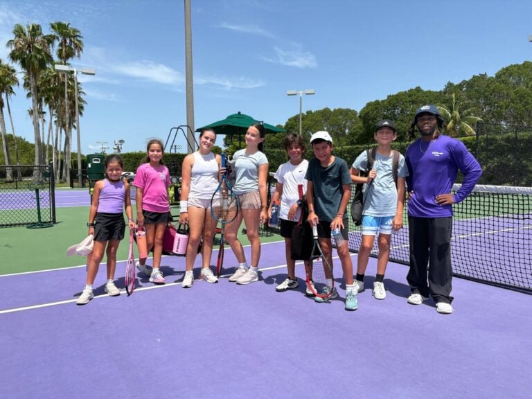 Eight people, seven teens and one adult, stand on a purple outdoor tennis court holding rackets, smiling at the camera on a sunny day.