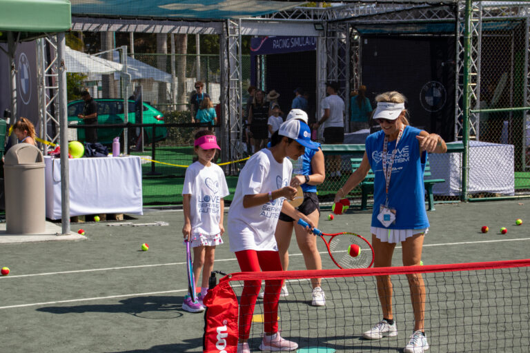 A tennis instructor in blue guides young players on a court during a lesson. Tennis balls and equipment are visible around the area.
