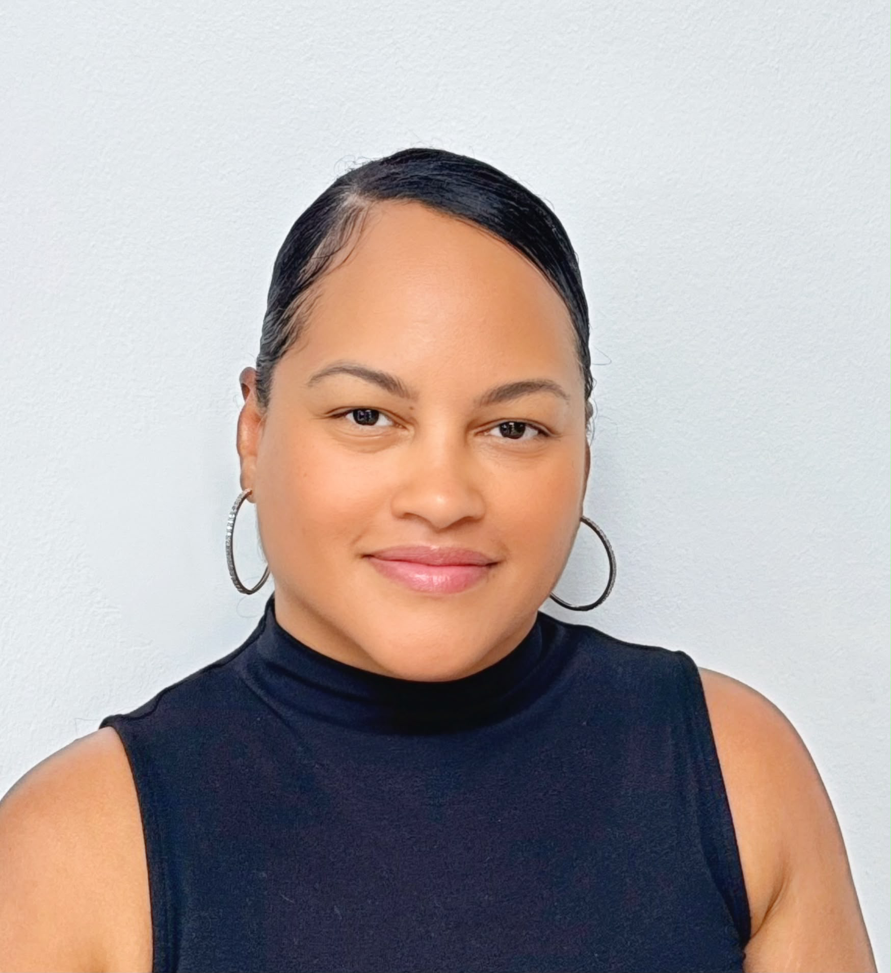 Woman with dark hair pulled back, wearing hoop earrings and a sleeveless black turtleneck top, smiles at the camera against a plain light background.