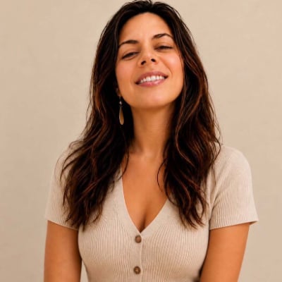 Woman with long brown hair wearing a beige ribbed top with buttons, standing against a plain light background, smiling with eyes partially closed.