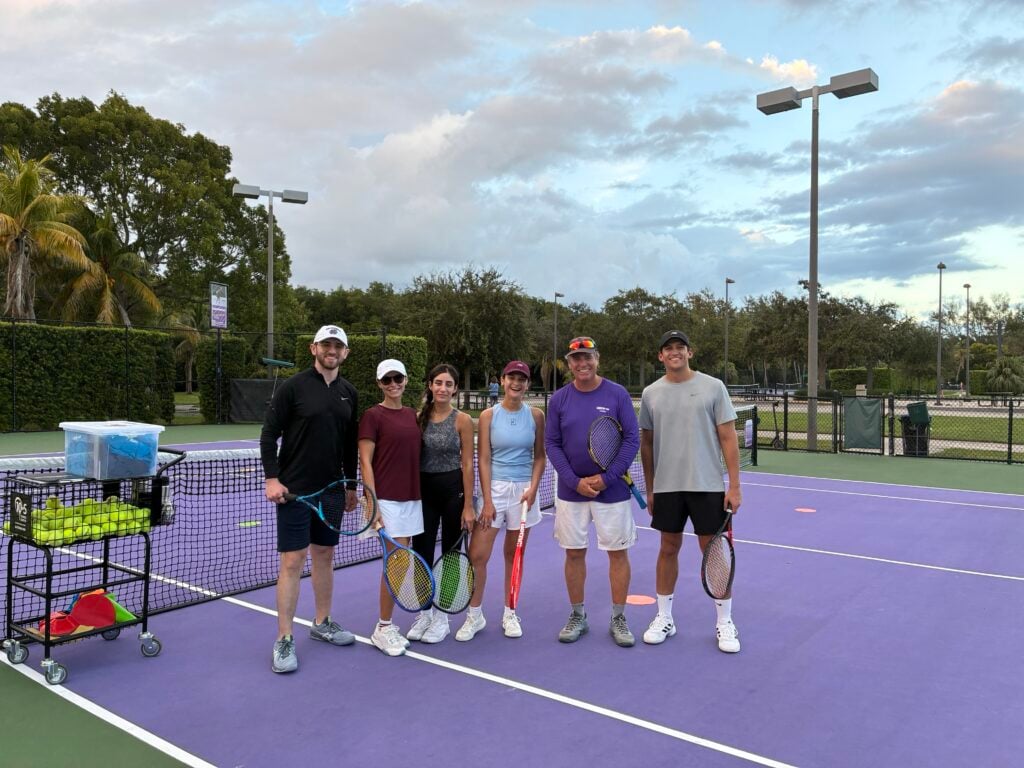 Six people stand on a purple tennis court holding rackets, with a cart of tennis balls nearby.