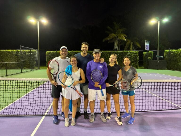 Six people standing on a tennis court at night, holding tennis rackets.
