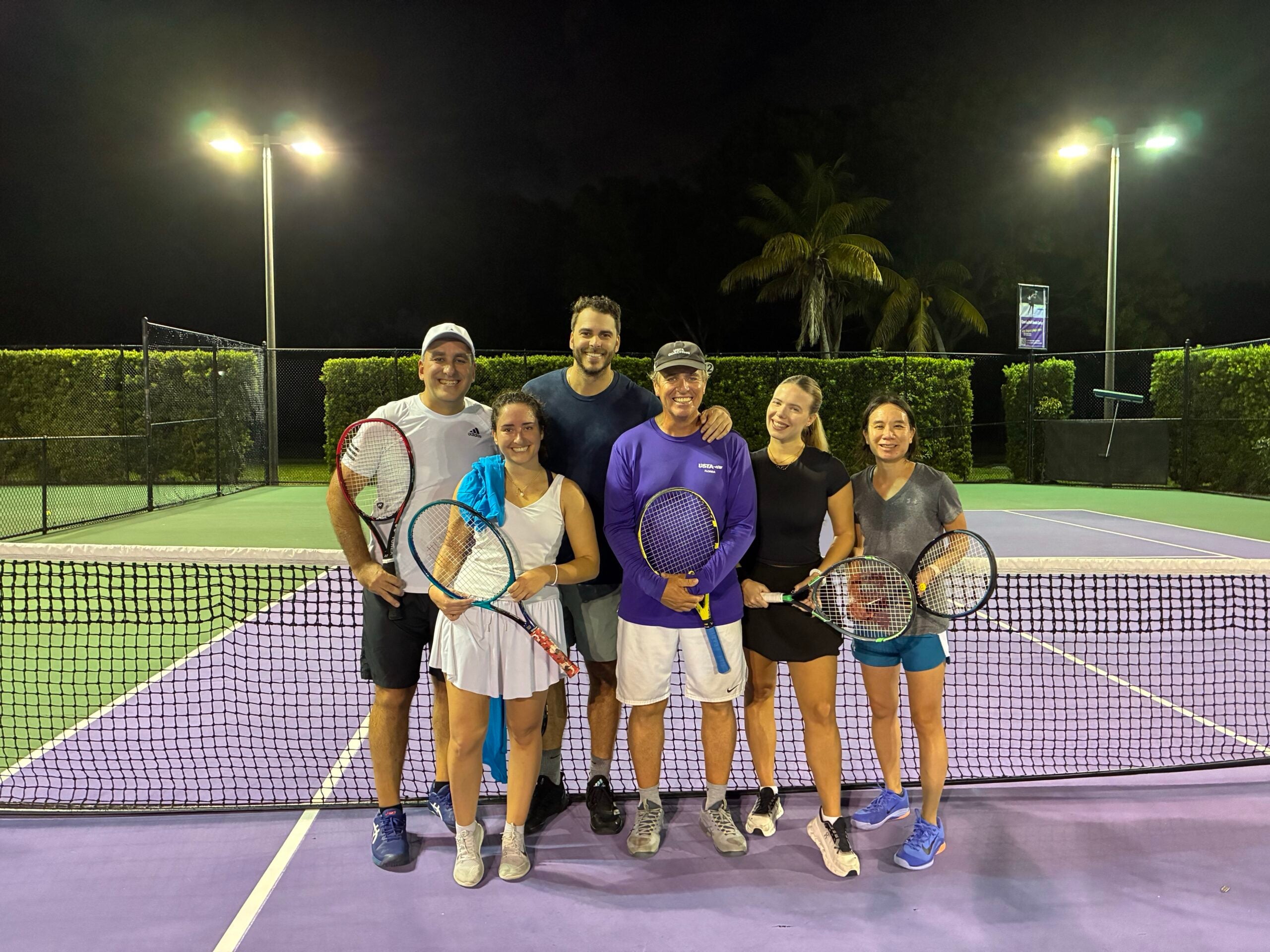 Six people standing on a tennis court at night, holding tennis rackets.